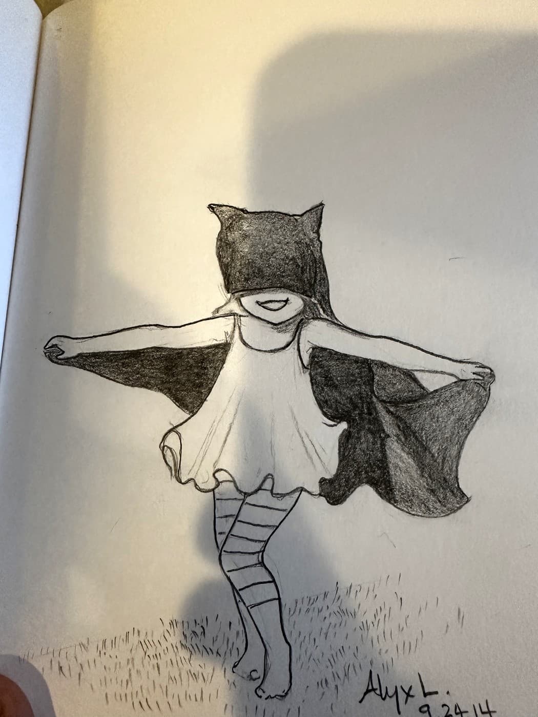 A pencil drawing in a sketchbook of a girl in a pointed-eared mask and a dark cape, arms out, spinning in a field of drawn grass. Signed Alyx L. and dated 9.24.14.